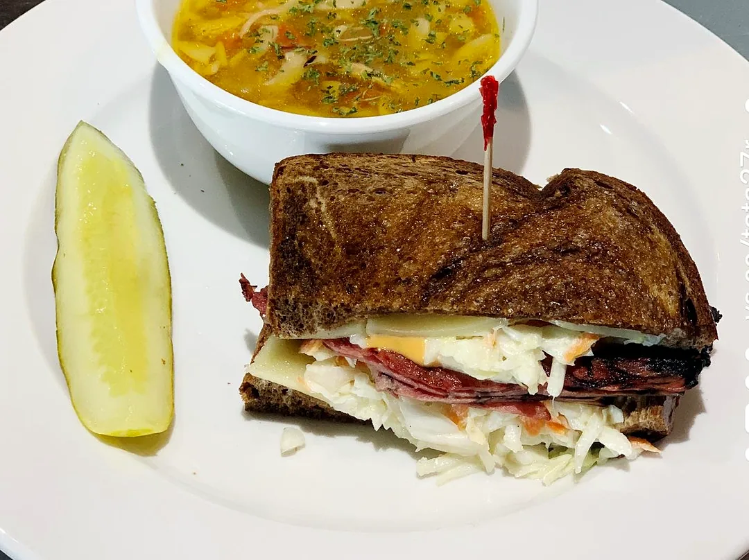 A grilled reuben with a cup of soup and a pickle
